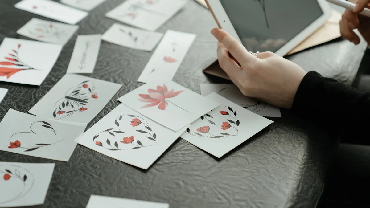A person drawing floral artwork on a tablet beside printed floral designs